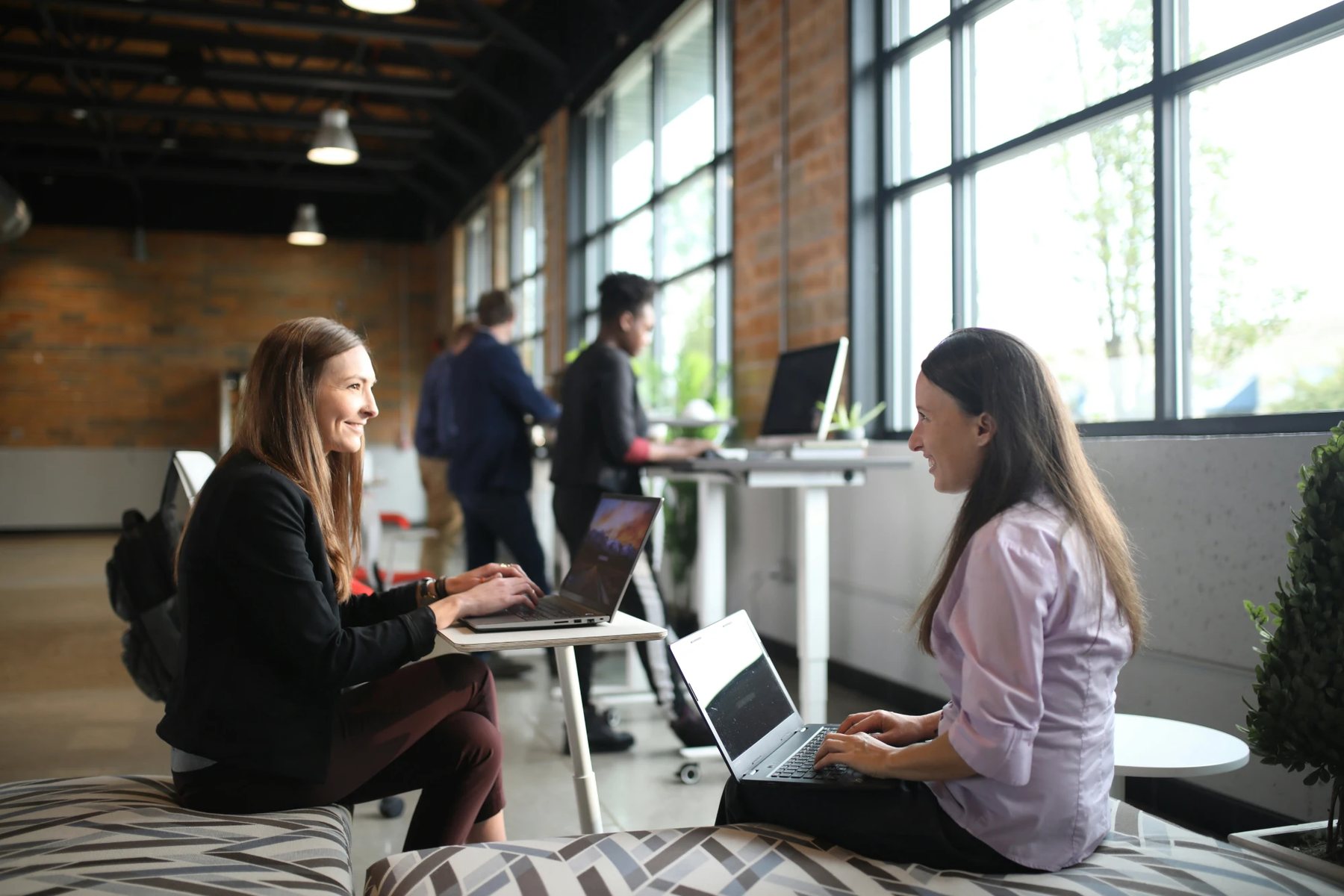 Two colleagues working on laptops in an open office, talking as they work